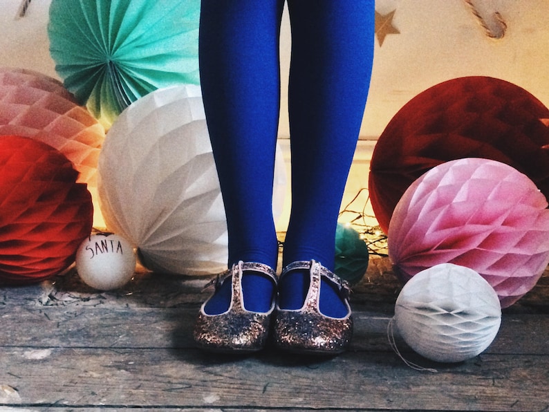 May include: A festive scene featuring blue tights and glittery gold shoes with T-straps. The background includes colorful paper honeycomb decorations in red, pink, white, and teal, with a small ornament that says "SANTA".
