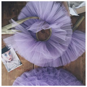 Op de afbeelding: Een close-up van drie lavendelkleurige tule tutu's met gouden elastische taillebanden. Links een kleine foto van een persoon in clownskostuum. De tutu's liggen op een houten oppervlak.