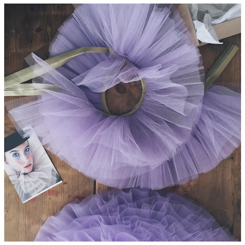 Op de afbeelding: Een close-up van drie lavendelkleurige tule tutu's met gouden elastische taillebanden. Links een kleine foto van een persoon in clownskostuum. De tutu's liggen op een houten oppervlak.