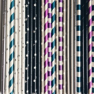 May include: A close-up of a variety of patterned paper straws. The straws are arranged in a row and feature different colors and patterns, including black and white stars, purple and white stripes, and teal and white stripes.