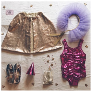 Op de afbeelding: Een verzameling kinderfeestkleding en accessoires. Inclusief een gouden pailletten cape, een paarse tule hoofdband, een roze metallic badpak, glitterschoenen en een gestreepte zak. Gouden sterrenconfetti en een roze feesthoed maken de feestelijke sc&egrave;ne compleet.