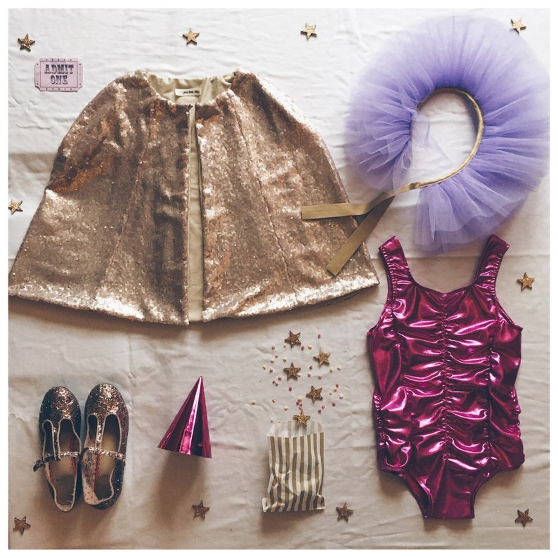 Op de afbeelding: Een verzameling kinderfeestkleding en accessoires. Inclusief een gouden pailletten cape, een paarse tule hoofdband, een roze metallic badpak, glitterschoenen en een gestreepte zak. Gouden sterrenconfetti en een roze feesthoed maken de feestelijke sc&egrave;ne compleet.