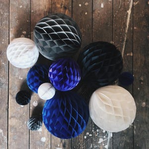 May include: An assortment of decorative paper honeycomb balls in various sizes and colors, including white, black, and shades of blue. The balls are arranged on a rustic wooden surface, creating a visually appealing display for party decorations.