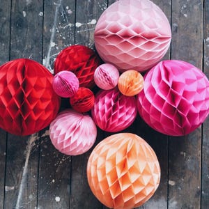 May include: A collection of colorful paper honeycomb balls in shades of red, pink, and orange. The decorative spheres vary in size and are arranged on a weathered wooden surface, creating a vibrant and festive display.