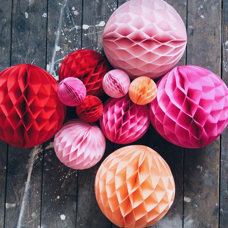 May include: A collection of colorful paper honeycomb balls in shades of red, pink, and orange. The decorative spheres vary in size and are arranged on a weathered wooden surface, creating a vibrant and festive display.
