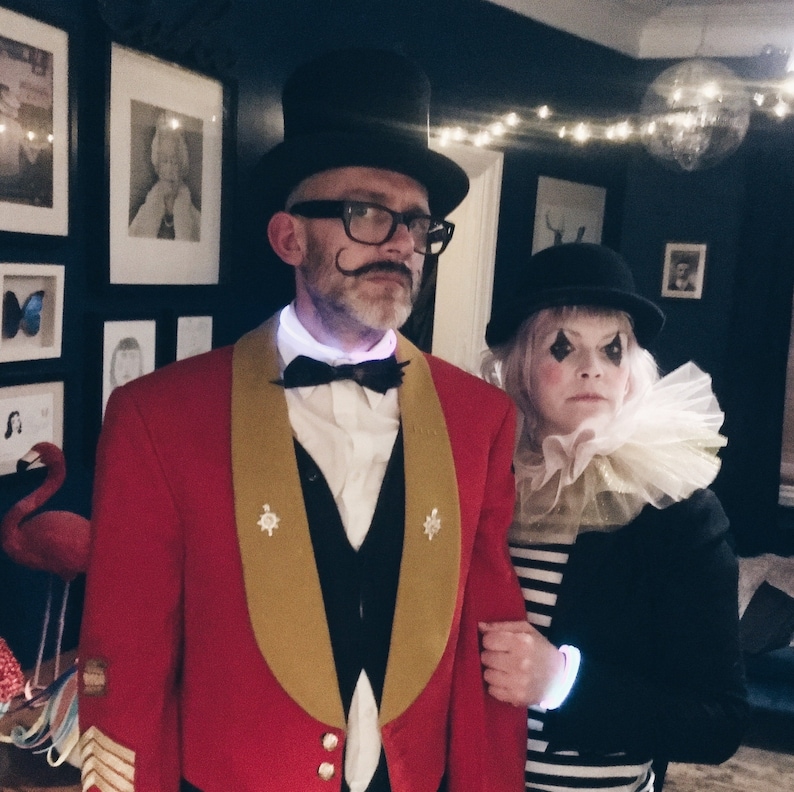 Op de afbeelding: Een stel in kostuums, gekleed voor een circus- of theatervoorstelling. De man draagt een rode jas met gouden rand, een hoge zwarte hoed en een vlinderdas. De vrouw draagt een zwarte bolhoed, een geplooide kraag en een gestreept shirt.