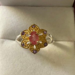 May include: A silver ring featuring a pink oval gemstone set in a gold flower design with purple accents.