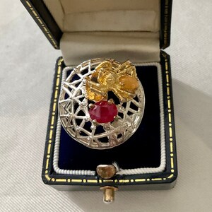 May include: A silver ring featuring a red gemstone and a gold bee design. The ring is presented in a black velvet box.