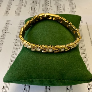 Peut inclure: Un bracelet en or avec un design délicat orné de petites pierres claires. Le bracelet est présenté sur un coussin de velours vert.