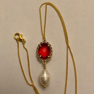Peut inclure: Un collier doré orné d'un pendentif en forme de pierre précieuse rouge et d'une perle blanche en forme de goutte. La pierre rouge est ovale et sertie dans un cadre doré décoratif. La chaîne est fine et dorée.