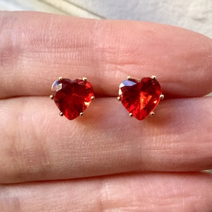Peut inclure: Une paire de boucles d'oreilles à clous en forme de cœur. Chaque boucle d'oreille présente une pierre précieuse rouge vif à facettes sertie dans un métal de couleur or. Les pierres précieuses sont taillées pour maximiser la réflexion de la lumière.