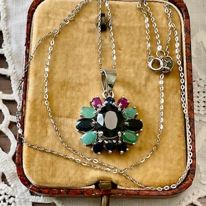 Peut inclure: Un collier en argent avec un pendentif orné d'une grosse pierre sombre entourée de petites pierres précieuses colorées. Le pendentif est serti dans un motif floral avec des pierres vert émeraude, noires et couleur rubis. Le collier est présenté dans un écrin vintage.
