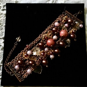 Peut inclure: Bracelet à plusieurs rangs avec une base en chaîne de bronze. Le bracelet est orné de perles marron, roses et transparentes de différentes tailles. Les perles sont regroupées, créant un aspect superposé et texturé. Le bracelet est présenté sur une surface noire.