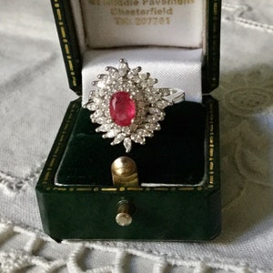 Può includere: Un anello con rubino rosso incastonato in una fascia d'argento con un gruppo di piccoli diamanti attorno al rubino. L'anello è in una scatola per anelli in velluto verde.