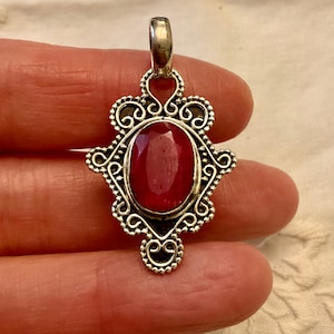 May include: A silver pendant with an ornate design featuring a red gemstone in the centre.