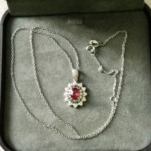 Peut inclure: Un collier en chaîne argent avec un pendentif orné d'une grande pierre rouge ovale entourée de petites pierres claires.