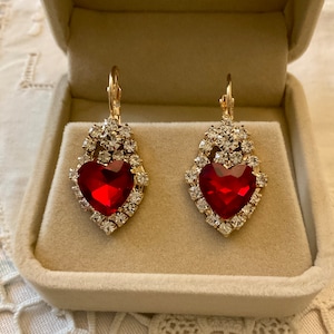 Peut inclure: Paire de boucles d'oreilles dorées avec une pierre centrale rouge rubis en forme de cœur. Les boucles d'oreilles sont entourées de cristaux clairs et scintillants, présentées dans un écrin beige.