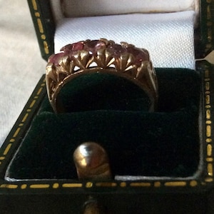 May include: A gold ring with a row of pink gemstones set in a decorative design. The ring is in a green velvet ring box.
