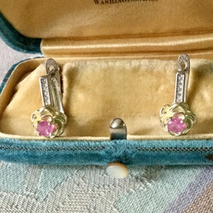 Peut inclure: Boucles d'oreilles en argent avec des pierres roses et de petits brillants, présentées dans un écrin en velours bleu à l'intérieur doré. Les boucles d'oreilles ont un motif floral.