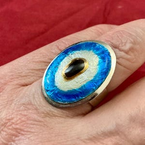 Può includere: Un anello ovale caratterizzato da una forma centrale nera allungata circondata da un anello bianco e da un bordo esterno blu testurizzato. Il design è incastonato in una montatura in metallo argentato, creando un aspetto stratificato sul gioiello.