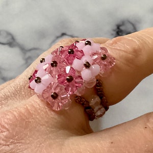 Peut inclure: Une bague en perles roses et blanches avec un motif floral. La bague est faite de petites perles rondes qui sont enfilées ensemble pour créer un motif floral. La bague est réglable et peut être portée à n'importe quel doigt.