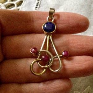 Op de afbeelding: Een zilveren en gouden hanger met een blauwe edelsteen en drie roze edelstenen. De hanger heeft de vorm van een gestileerde boom met een enkele tak die zich vanaf de bovenkant uitstrekt.