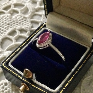 May include: A silver ring with a pink marquise-shaped gemstone surrounded by smaller diamonds. The ring is in a blue velvet ring box.