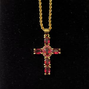 Könnte beinhalten: Eine goldfarbene Halskette mit einem Kreuzanhänger. Das Kreuz ist mit ovalen roten Edelsteinen und kleineren roten Steinen besetzt. Die Kette hat ein gedrehtes Seildesign. Der Hintergrund ist schwarz.