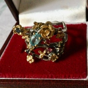 Peut inclure: Une bague en argent avec un motif floral orné de pierres précieuses bleues, rouges et dorées. La bague est placée dans une boîte avec un revêtement en velours rouge.