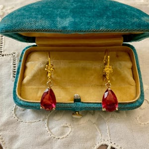 Peut inclure: Une paire de boucles d'oreilles pendantes dorées avec des pierres rouges facettées en forme de poire, suspendues à de petits ornements métalliques en forme de fleur. Elles sont présentées dans un écrin en velours bleu sarcelle à intérieur jaune.