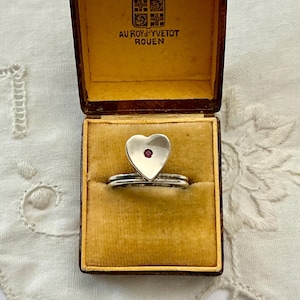 Peut inclure: Une bague en argent avec une face en forme de cœur et une petite pierre précieuse rouge au centre, présentée dans un coffret vintage marron avec un intérieur doré. La boîte est ouverte, révélant la bague.