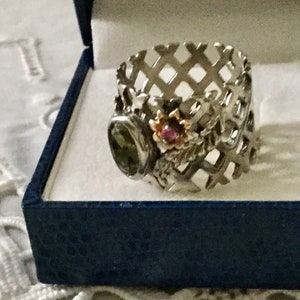 Peut inclure: Une bague en argent avec une pierre précieuse verte, une pierre précieuse rose et un motif de fleur en or. La bague est sertie dans un motif de treillis en argent.