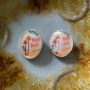 Peut inclure: Deux pendentifs de charme argentés avec un fond blanc et des palmiers. Le texte "Miami Beach" est imprimé en rose sur chaque charme. Chaque charme est décoré de strass turquoise.