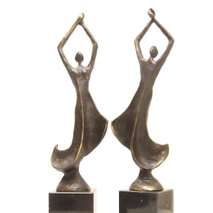 Può includere: Due sculture in bronzo di figure in abiti fluenti con le braccia alzate. Ogni figura è montata su una base quadrata nera. Le sculture hanno un design classico ed elegante, adatto all'arredamento della casa.