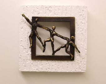Op de afbeelding: Een bronzen sculptuur van vier figuren die elkaar de hand reiken, ingelijst in een donkerbruin frame met een witte textuur achtergrond.