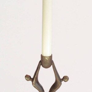 Può includere: Un portacandele color bronzo con una candela bianca. Il supporto ha un design unico con due figure che sostengono la coppa della candela. La base è quadrata e l'altezza è di 21 cm.