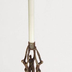 Può includere: Un portacandele color bronzo con tre figure umane stilizzate che sostengono una candela bianca. Le figure sono in posa dinamica, con le braccia alzate. La candela è alta e cilindrica. La base è triangolare. Altezza: 20 cm.