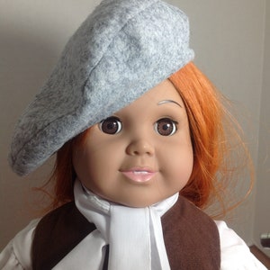 May include: A doll with red hair wearing a grey beret and a white scarf over a brown waistcoat.
