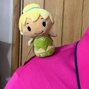 Peut inclure: Une poupée en peluche de fée avec des cheveux jaunes et une robe verte. La poupée est posée sur un polo rose vif avec une bordure bleu marine. La fée a des yeux noirs et un petit sourire.