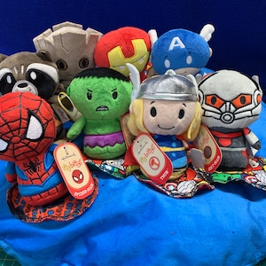 Puede incluir: Una colección de peluches con personajes del universo Marvel. Los peluches están dispuestos sobre una superficie de tela azul. Los personajes incluyen Spider-Man, Hulk, Thor, Ant-Man, Capitán América, Iron Man y Groot.