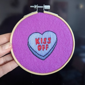 Peut inclure: Broderie en forme de bonbon en cœur avec le texte "KISS OFF" en lettres rouges sur fond bleu. La broderie est montée dans un cercle en bois avec un dos en feutre violet.