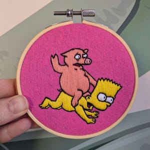 May include: Embroidered hoop art showing a pink pig riding a yellow cartoon character, set against a bright pink felt background. The wooden hoop holds the design, a fun, quirky piece.