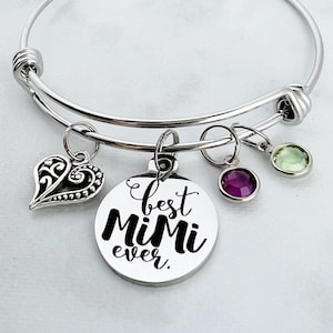 May include: A silver bangle bracelet with a charm that reads "best Mimi ever." The charm is attached to the bracelet with a silver ring. There are three small charms attached to the bracelet, one is a silver heart, one is a purple gemstone, and one is a green gemstone.