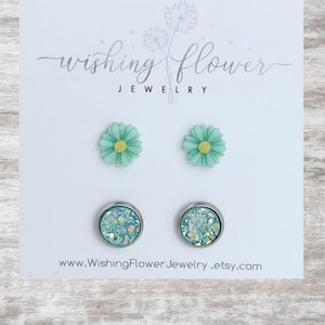 May include: A set of four earrings featuring two pairs of green flower stud earrings and two pairs of turquoise glitter stud earrings. The earrings are displayed on a white card with the text "Wishing Flower Jewelry" and a website address.