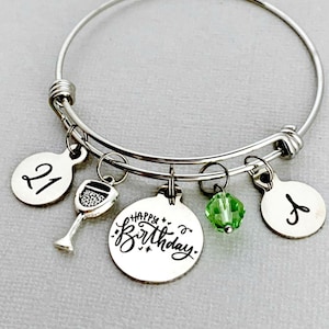 May include: A silver charm bracelet with a wine glass charm, a charm with the text "Happy Birthday", a green crystal charm, and two charms with the numbers "21" and "K" engraved on them.
