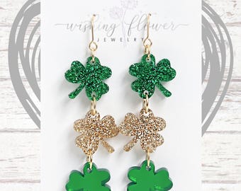 Könnte beinhalten: Ein Paar baumelnder Ohrringe mit St. Patrick's Day-Motiv. Jeder Ohrring hat drei Kleeblattformen aus grünem und goldfarbenem Glitzeracryl. Die Ohrringe sind an goldfarbenen Haken aufgehängt. Die Ohrringe werden auf einer weißen Karte mit dem Text "wishing flower JEWELRY" präsentiert.