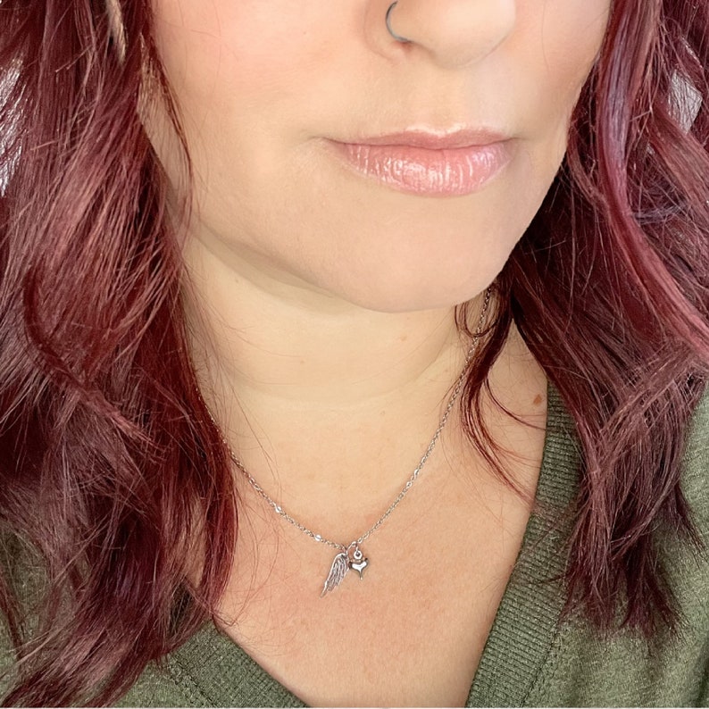 May include: A silver necklace with a small heart and angel wing charm. The necklace is worn by a person with red hair.