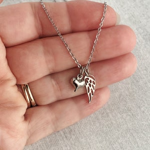 May include: A silver necklace with a small heart and angel wing charm. The necklace is on a hand.