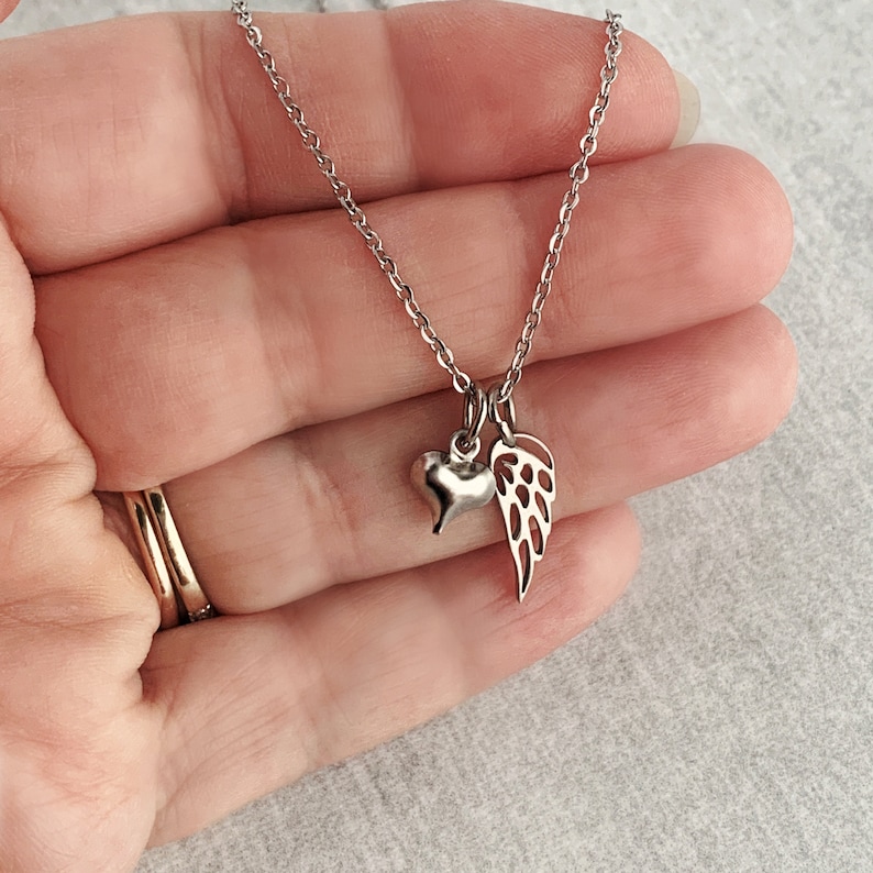 May include: A silver necklace with a small heart and angel wing charm. The necklace is on a hand.