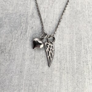 May include: A silver necklace with a heart and angel wing charm. The necklace is on a grey background.